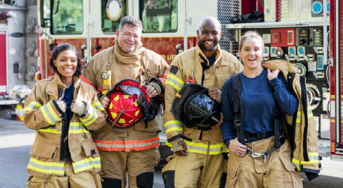 4 Firefighters standing next to a fire truck in full fire gear smiling