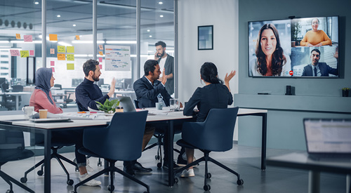 People sitting at a conference table looking toward a tv hanging on the wall with people joining their meeting virtually