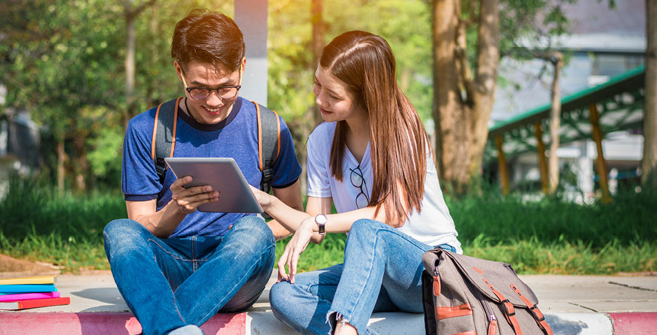 Two people sit next to each other on the curb while looking at a tablet.