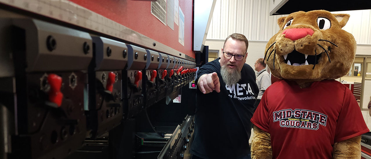 Mid-State mascot Grit standing next to an instructor pointing at some levers