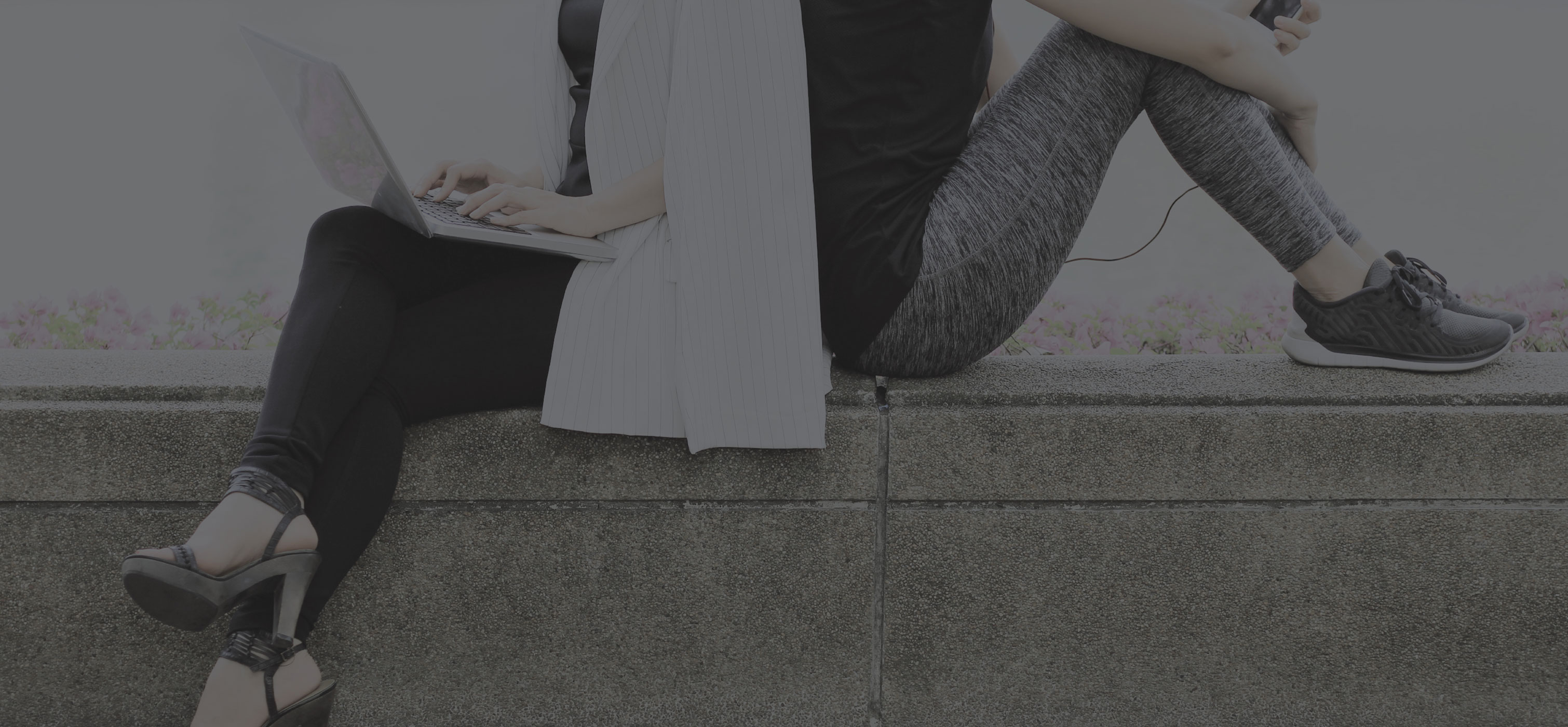 2 peole sitting on a wall with their backs facing each other. One dressed business casual holding a laptop the other dressed athletically holding a phone with headphones