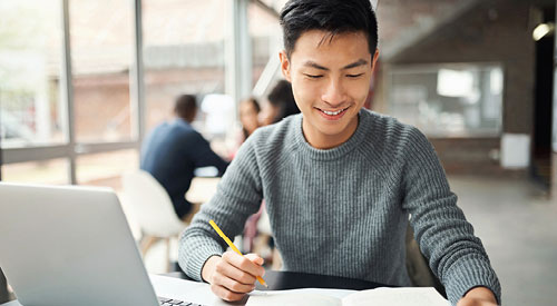 Person sitting in front of a laptop writing something down with a pencil