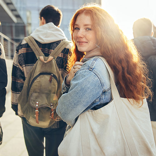 Group of 4 students with backpacks on walking toward a building. One student is turned around looking at the camera, sun shining in the background.