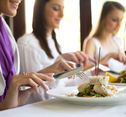 People sitting at a table with a white tablecloth holding knives and forks eating food off of white plates.