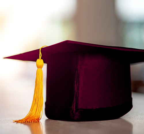 Maroon graduation cap with yellow tassel sitting on a table.