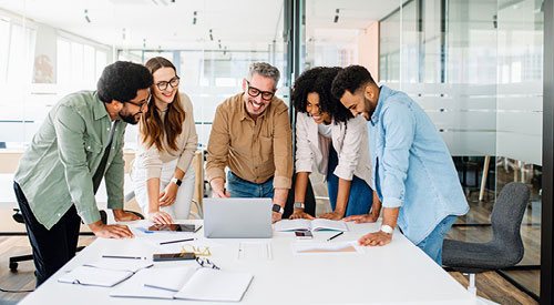 People in business attire standing in a circle talking around a table with a laptop and papers on it