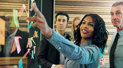 Person adding post it notes to a glass wall while others look on behind them