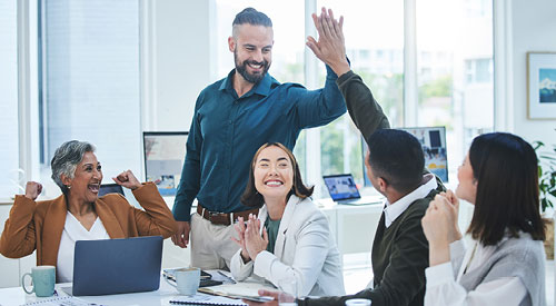 People sitting at a table with laptops and business supplies. 1 person is standing giving a high five to another person that is sitting