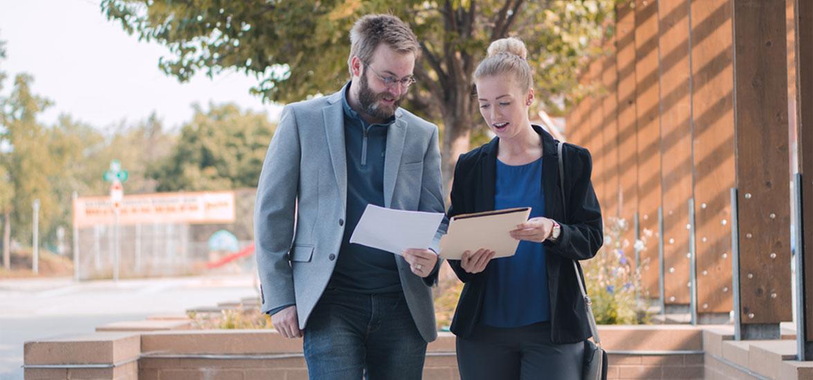 A man and woman dressed in semi-professional attire, holding papers and talking 