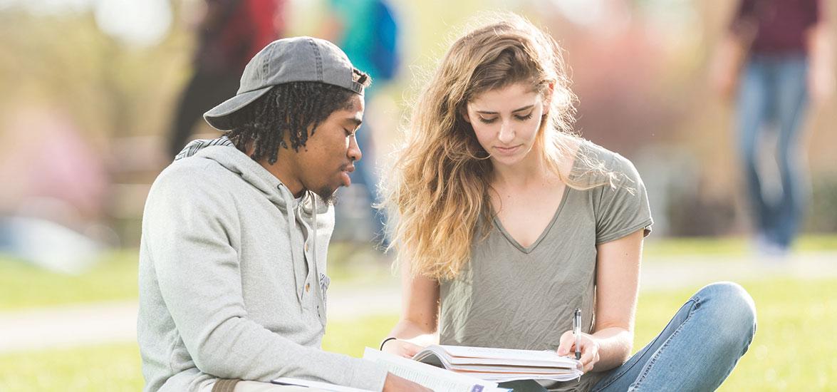 Two students, male and female, sitting on a lawn, reading and talking.