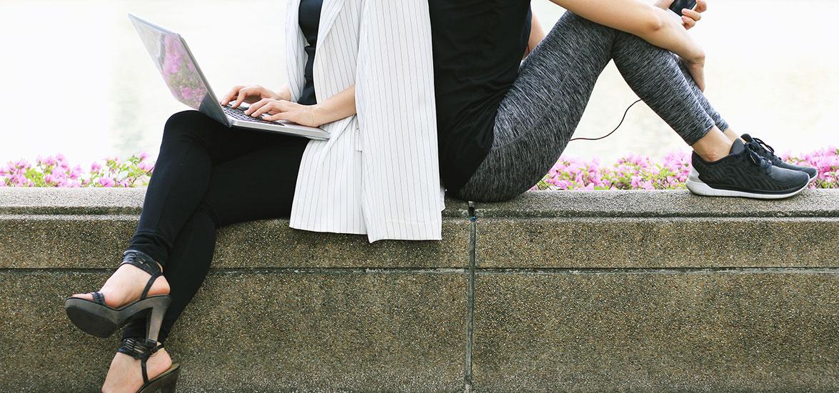 Two people sitting on a bench outside. One is in business clothes and one is in exercise clothes.